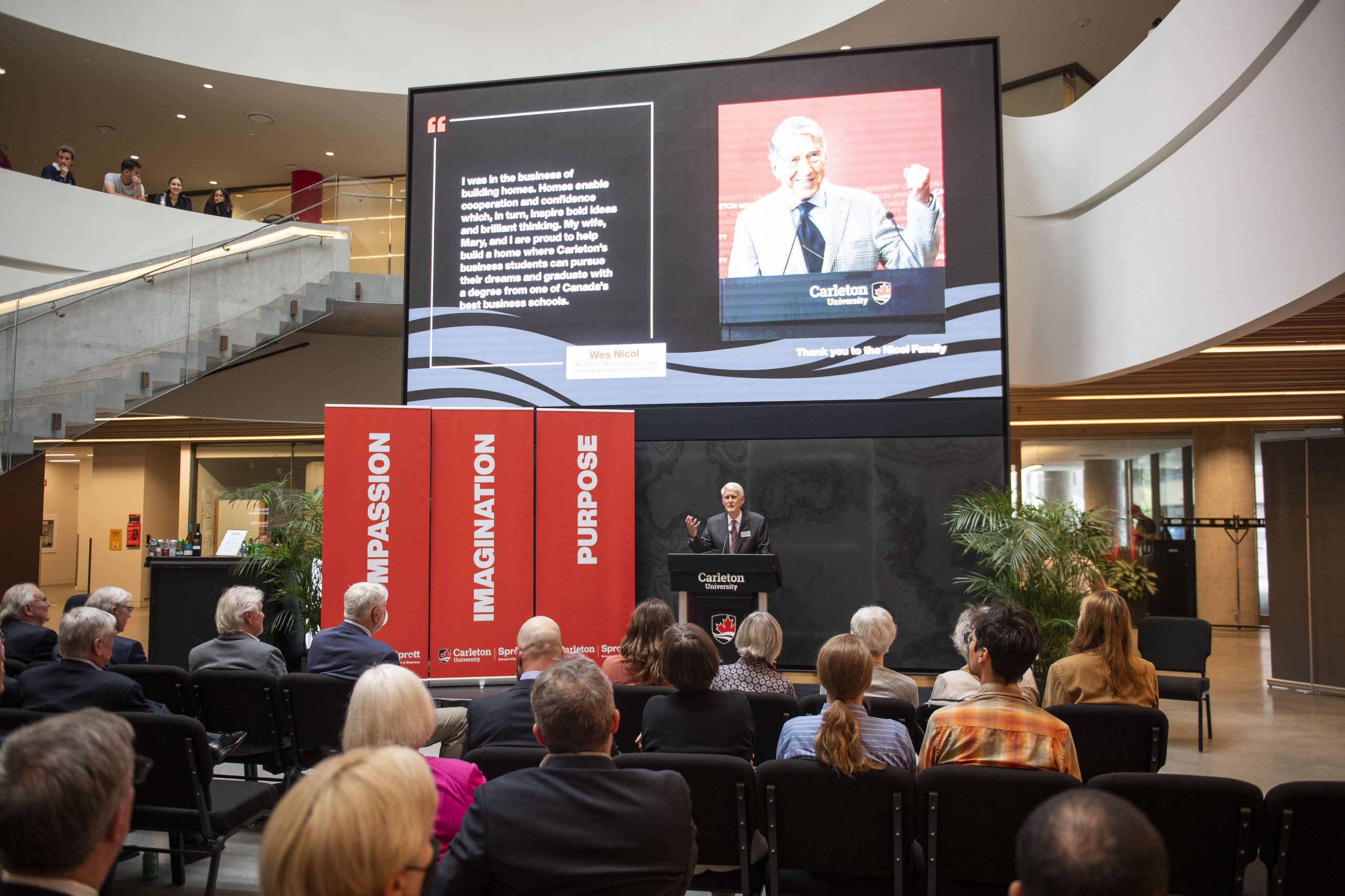 Carleton's Provost on stage in the atrium providing a speech in front of a screen with an image of Wes Nicol. 