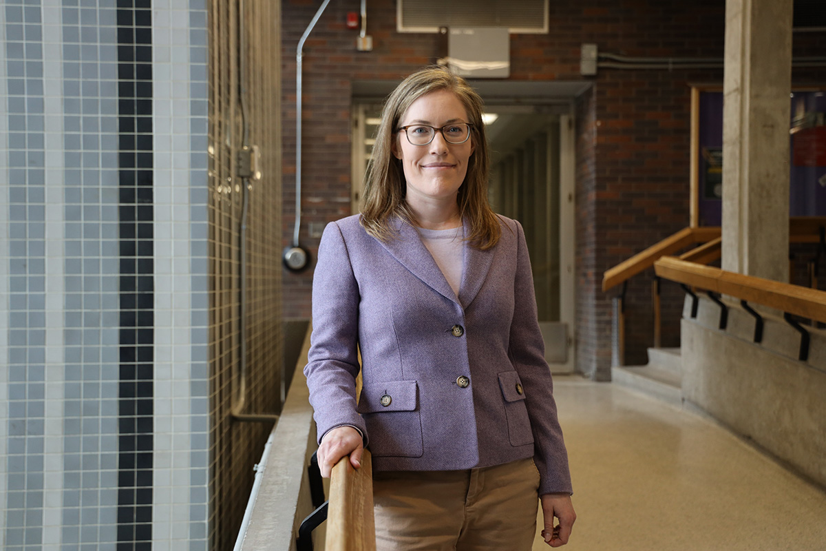 A woman wearing glasses leans against a railing inside a building to pose for a picture.