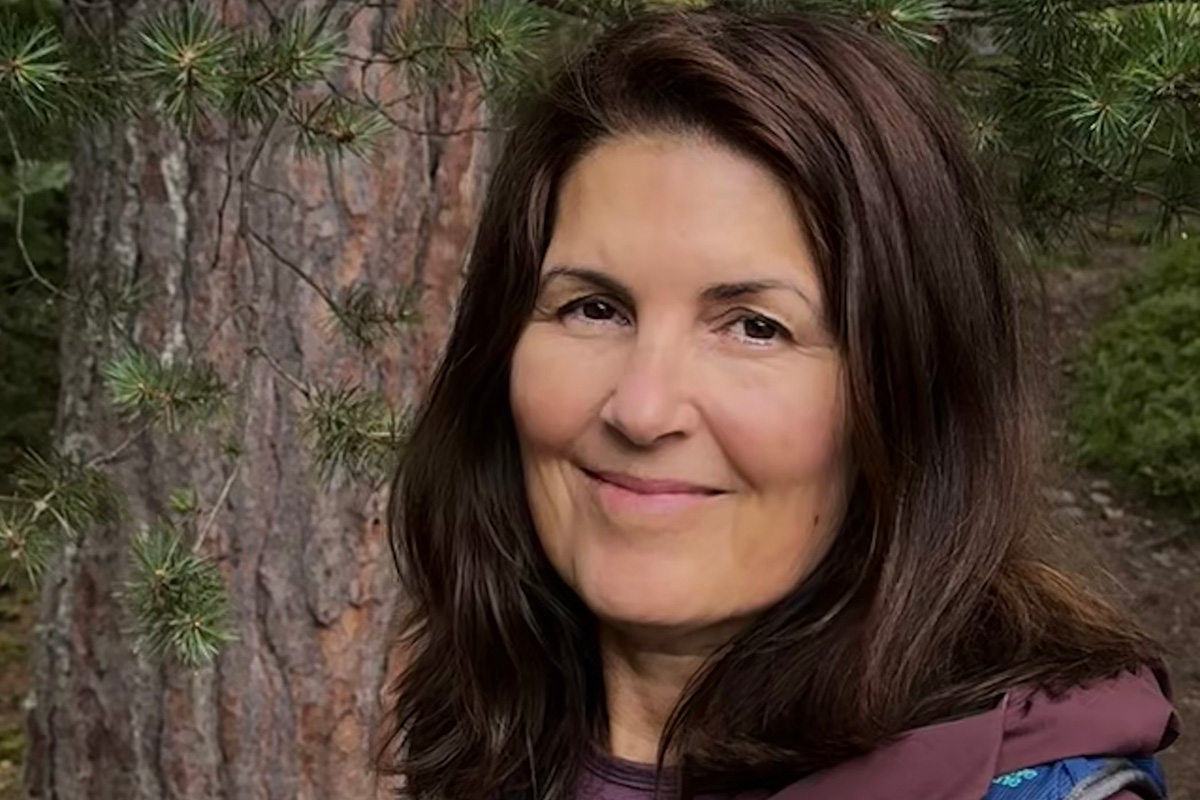 A woman with brown hair smiles for the camera while standing outside next to a tree.