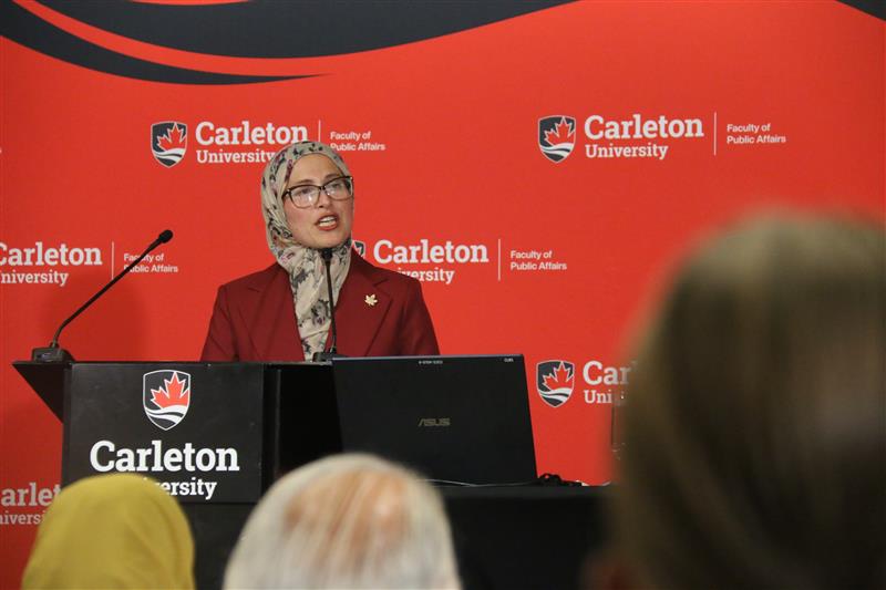 Woman wearing hijab speaking at a podium infront of a Carleton University banner. 