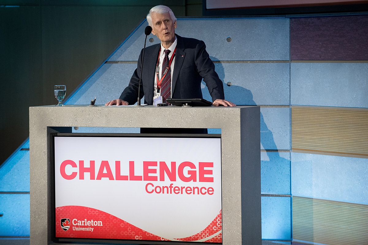 A man in a business suit speaks into a microphone while standing at a podium. The sign on the podium reads 'Challenge Conference' and includes the Carleton University logo.
