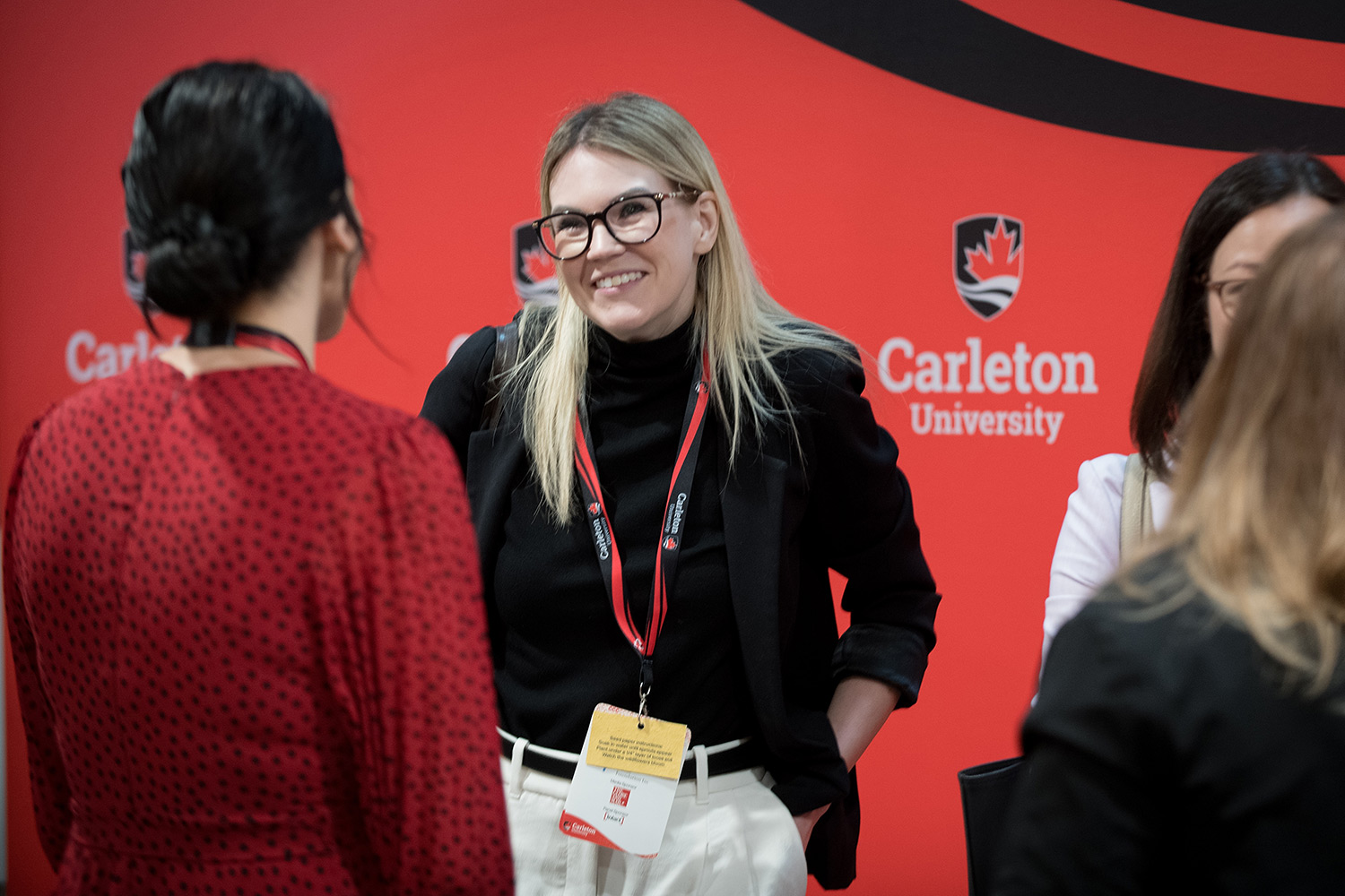 4 people engaging in conversations in front of a red promotional backdrop featuring the Carleton University logo.