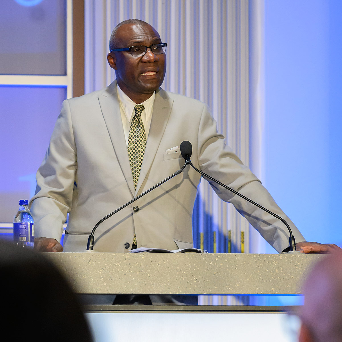 A man in a grey suit speaks into a microphone while standing at a podium at an AI conference.