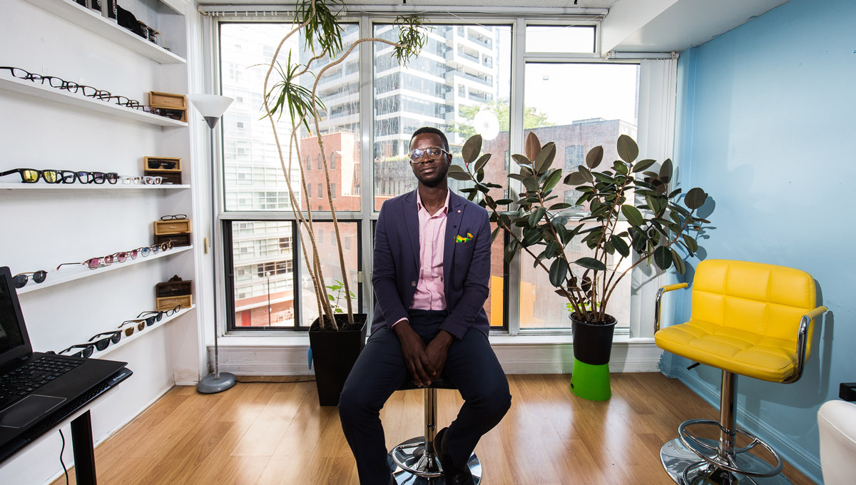 Nana Osei sit on a chair in his downtown office, with a wall of Bohten glasses and a plant in the background.