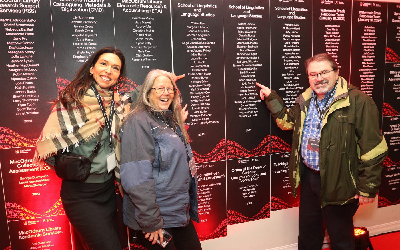 Three staff members pointing to their names on a wall of awards