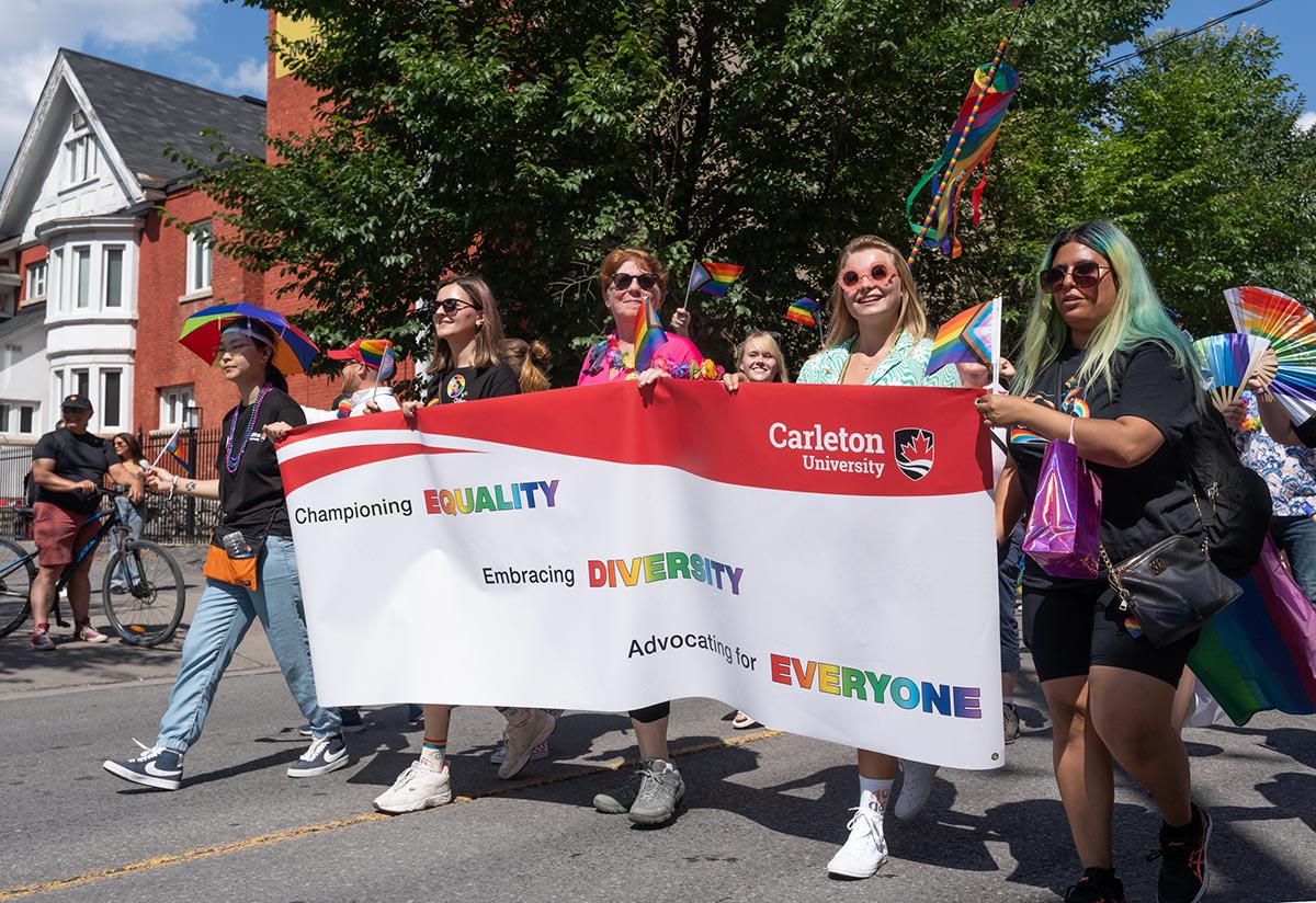 A group of people wearing colorful clothes hold a white and red sign as they march in the Pride parade