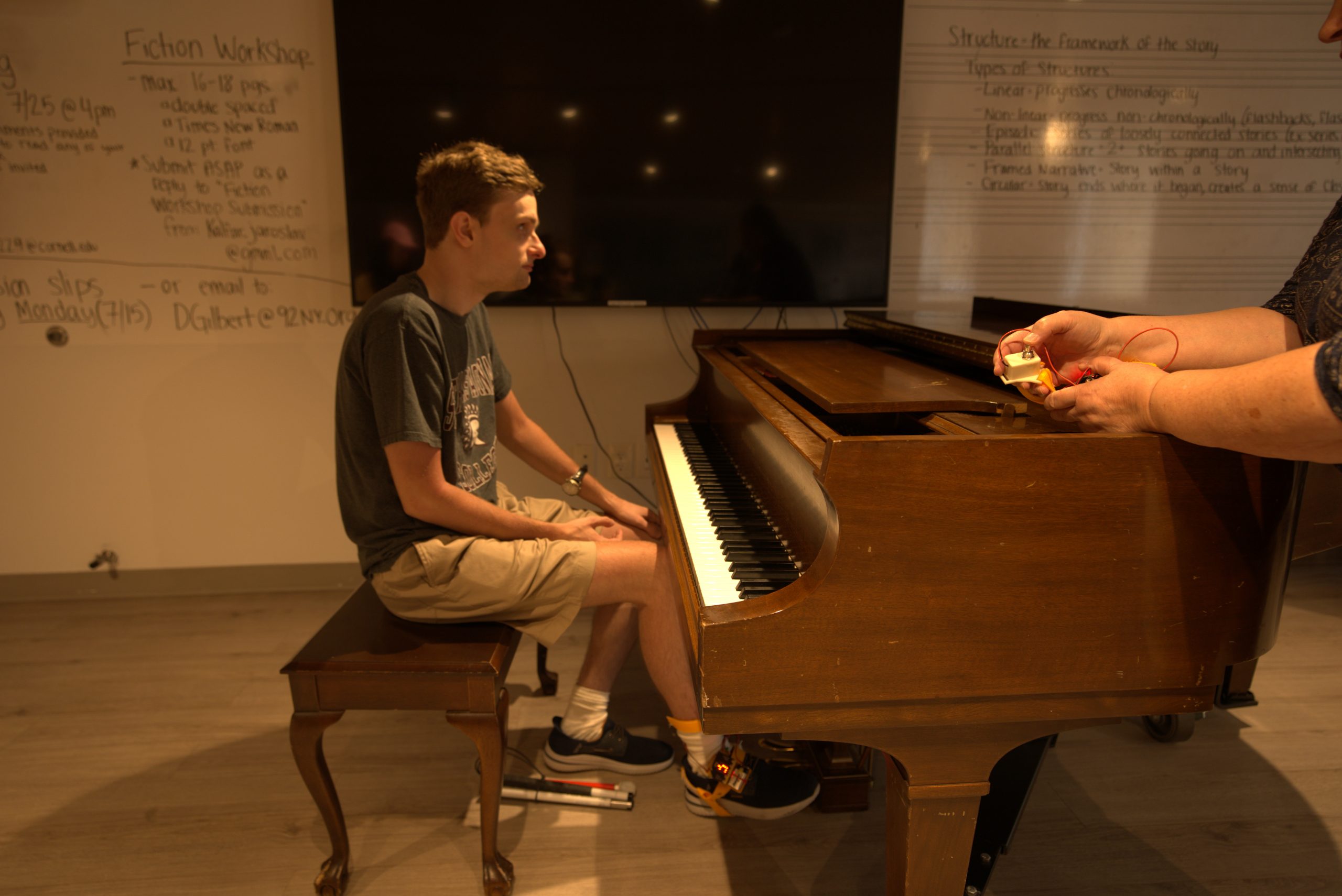 A man with light brown hair, a green shirt, and brown shorts, playing piano while wearing the tap-tap device around his ankle.