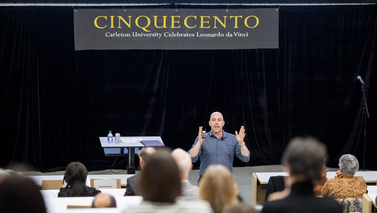 President Benoit-Antoine Bacon speaks about the 3D mastery of Da Vinci in front of an audience in the Health Sciences Building. A sign with the word Cinnquecento hangs above the podium.