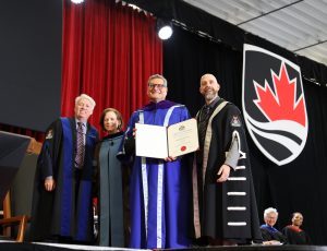 Four people stand on the stage at Carleton's Convocation and the one in the middle displays their degree