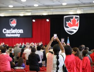 Attendees of Carleton's Convocation stand and watch the stage and one woman is taking a photo overhead with a cell phone