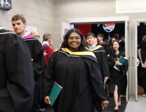 A group of graduates in black gowns walks out of Carleton's Fieldhouse and one woman smiles at the camera