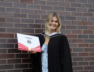 A women with blonde hair and a black Convocation gown smiles for the camera with her degree