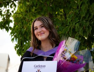 A graduating student poses with her degree and bouquet outside the Athletics Building.
