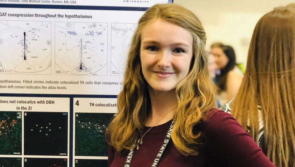 Kayla Schumacker stands in front of a poster board explaining her research.