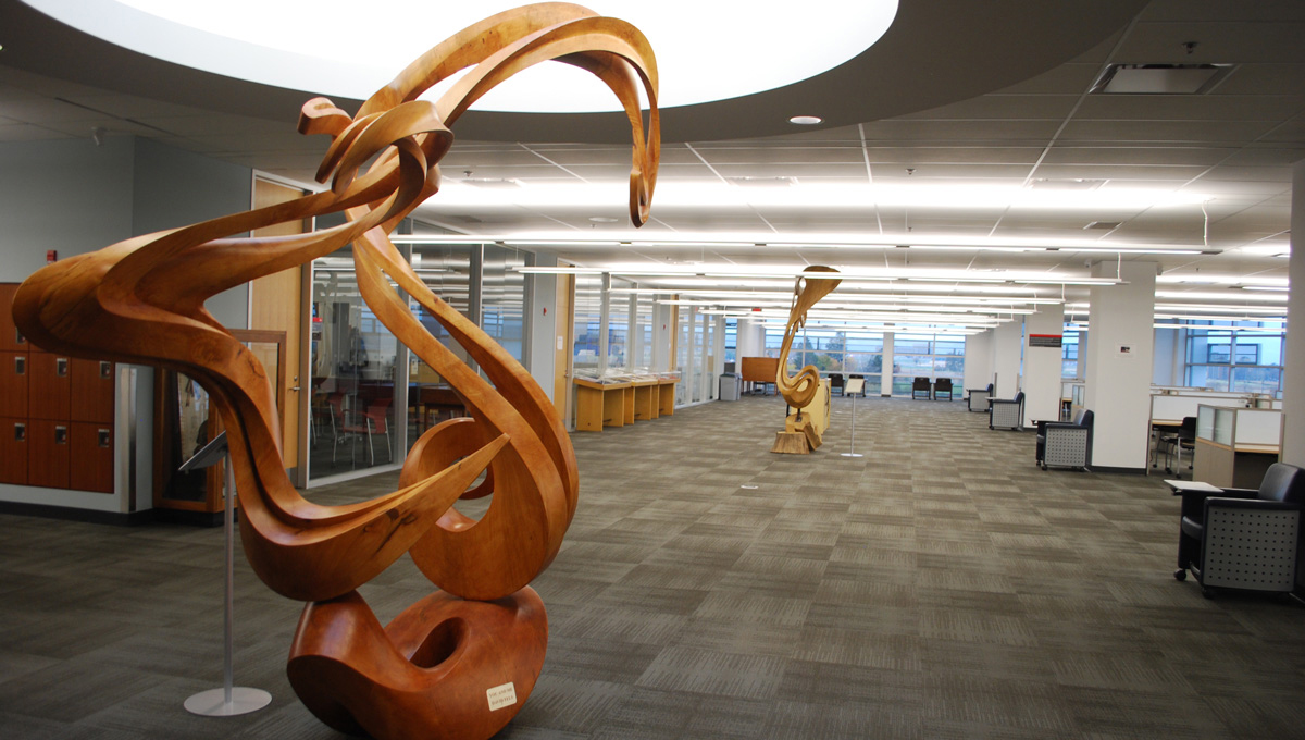 Wooden sculptures stand in a hallway in MacOdrum Library, glass-walled rooms are to the left of the sculpture and desks are on the right.