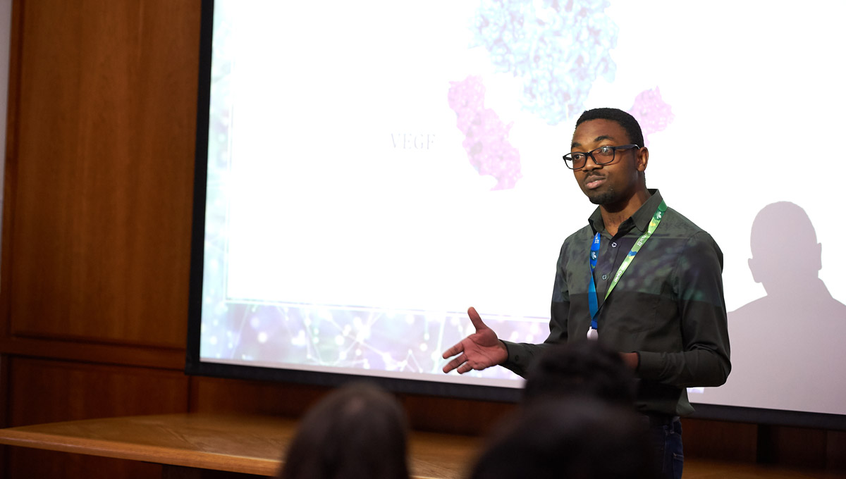 Panashe Kupakuwana speaks in front of a projector screen.