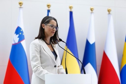 Woman in a white blazer giving a speech at a ing at a podium in front of a row of flags