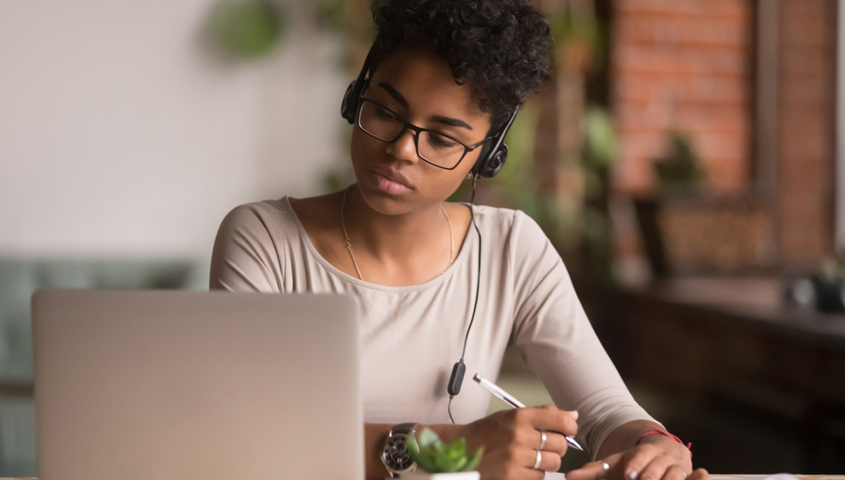 A woman wearing headphones and looking at her laptop computer takes notes