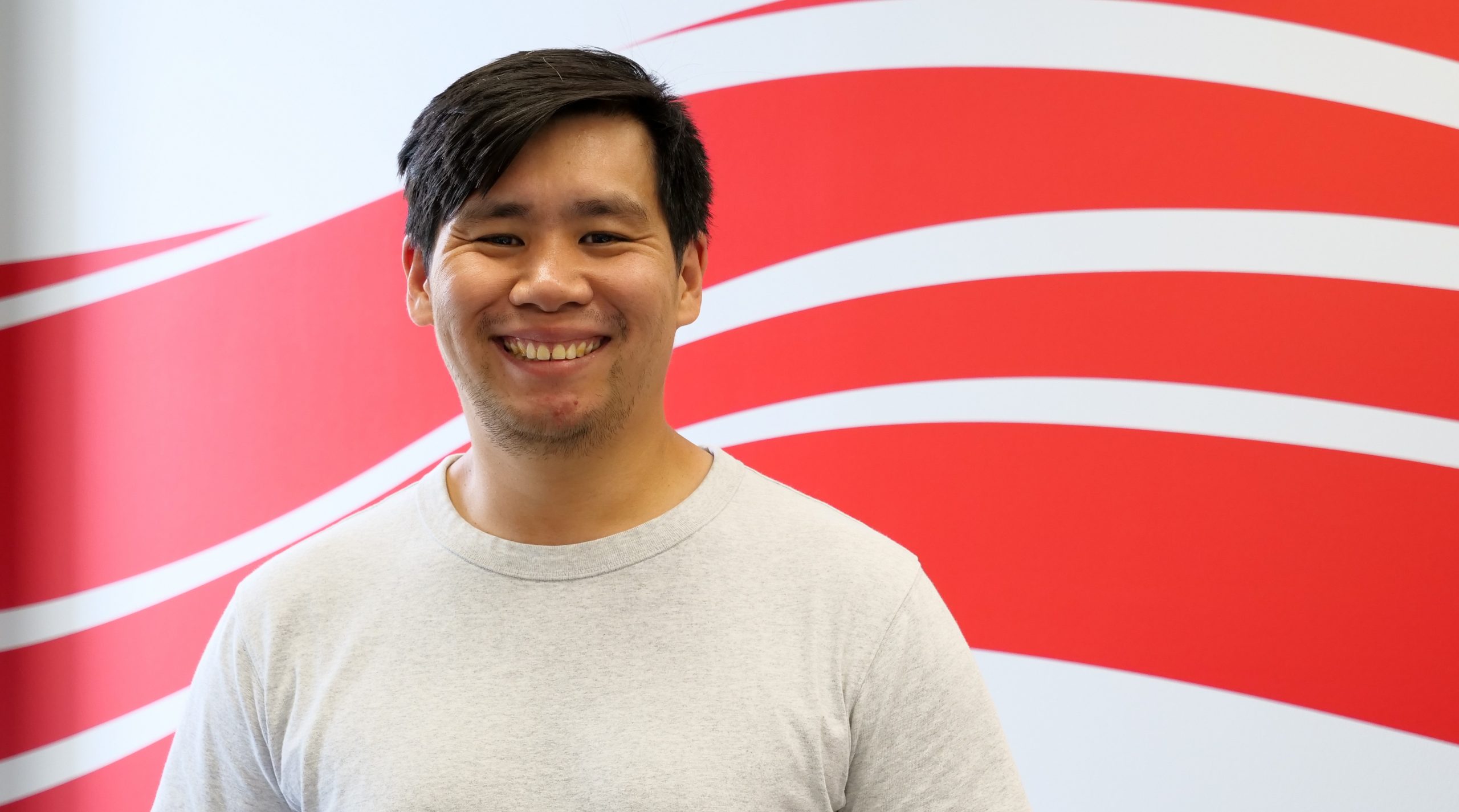 A man with black hair and light grey shirt in front of a white wall with a red swoop design.