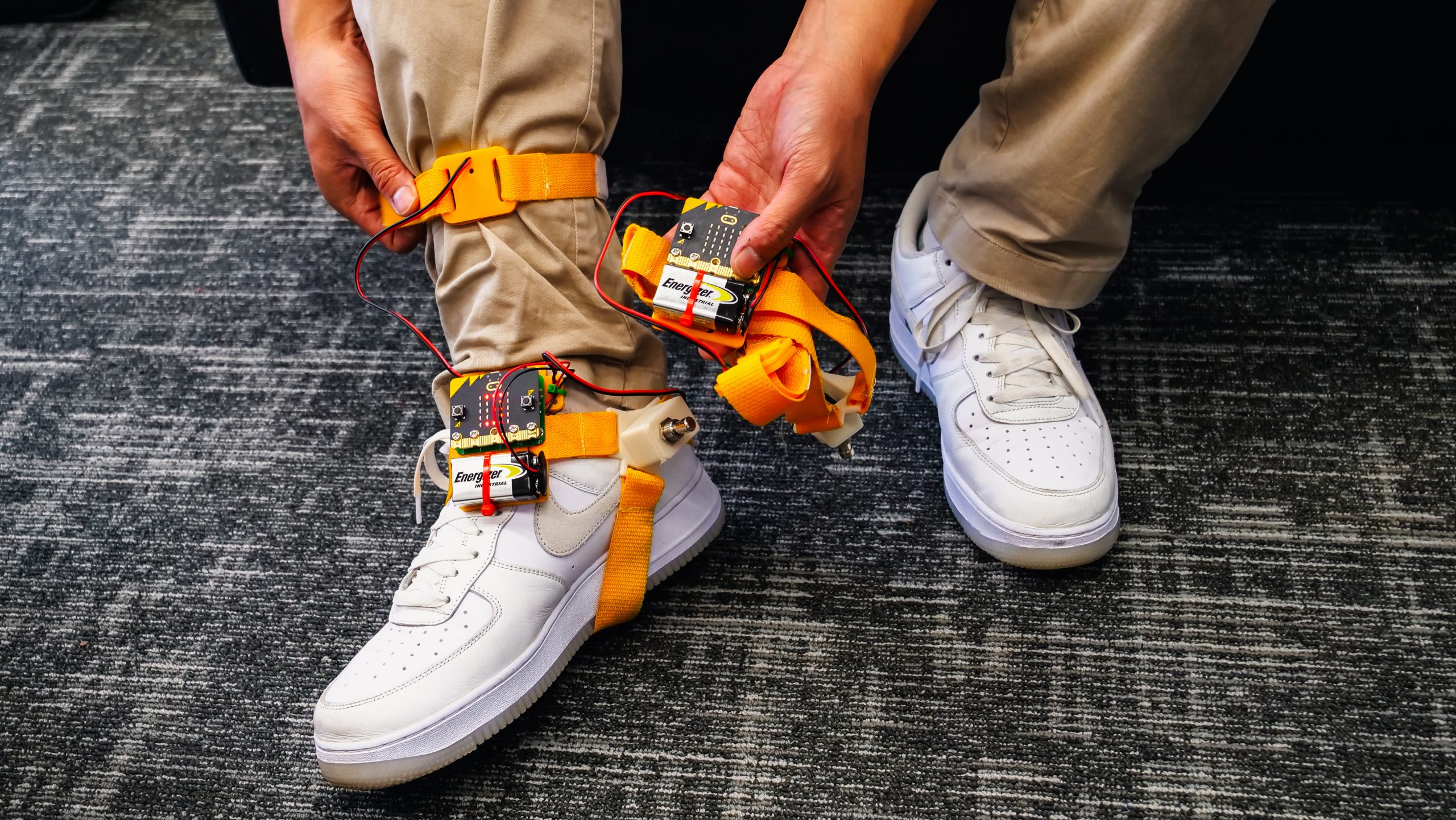 The tap-tap device, which is yellow and has a wires and circuit board, is strapped around someone wearing white Nike shoes.