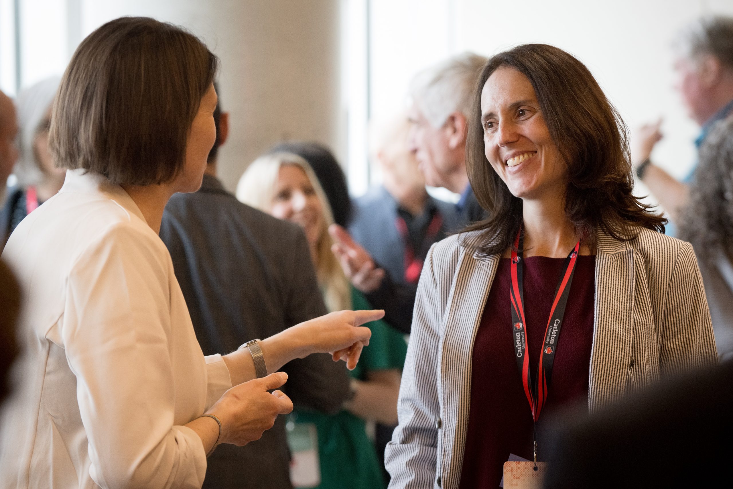 Two women networking at a conference event with a crowf of people behind them