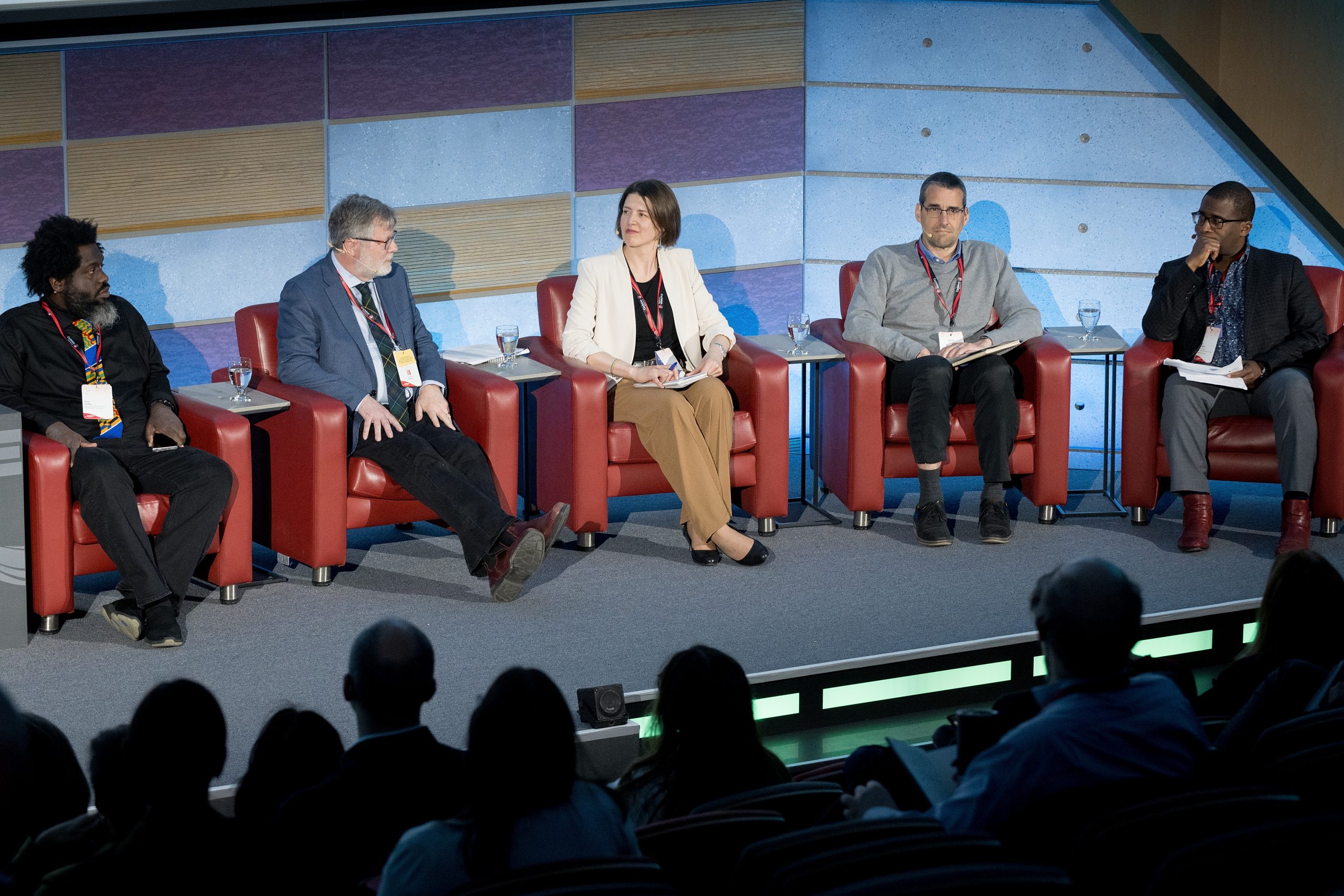 Five people on a conference panel engage in discussion in front of an audience.
