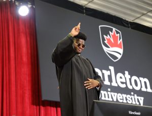 A male with sunglasses on holds his pointer finger to the sky on Carleton's Convocation stage