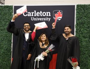 Three graduates pose in front of a Carleton branded banner while holding their degrees above their heads and smiling