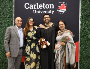 A Carleton graduate wearing a black down poses with his family in front of a Carleton branded banner
