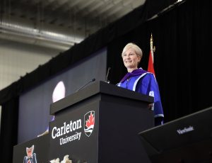A woman wearing a blue gown stands at the podium of Carleton's Convocation stage
