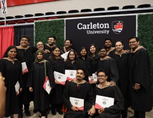 A large group of Carleton graduates wearing black gowns pose in front of Carleton's branded banner
