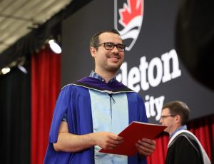 A Carleton graduate stands on the Convocation stage in a blue gown holding his degree