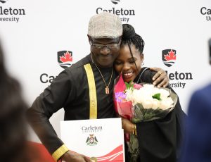 A Carleton graduate in a black gown holds flowers and hugs a family member