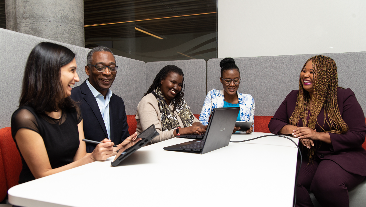 Group of smiling employees siting at a table with a computer and tablet to conduct work. 