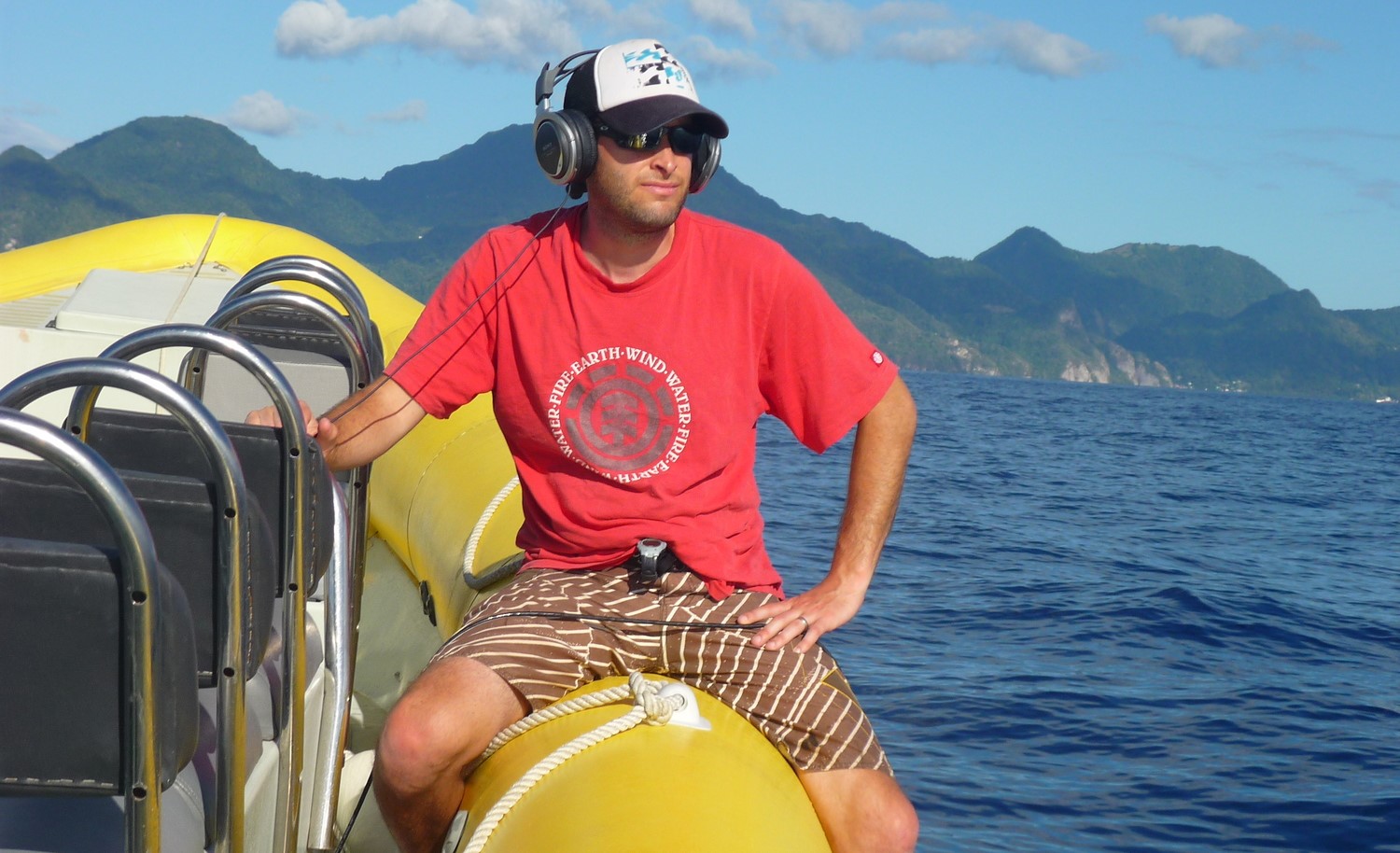 A man in a red shirt sits on a yellow boat with headphones on listening to whale communication