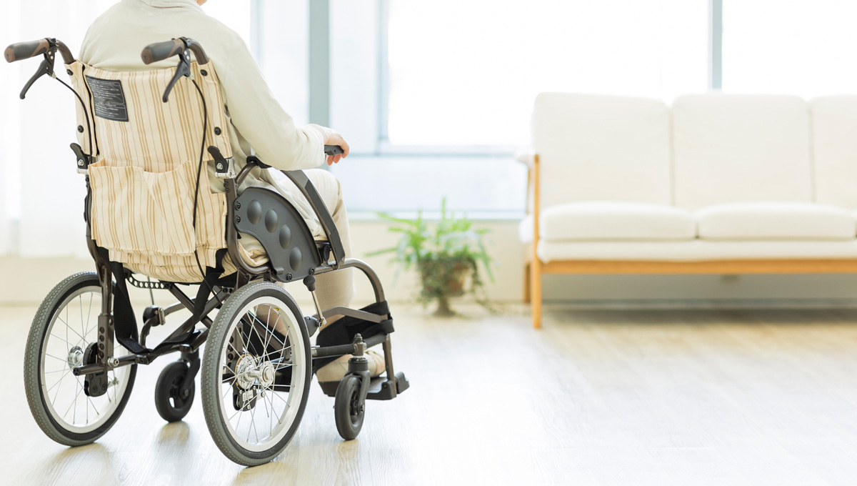 A woman sits in a wheelchair in a retirement home lounge. A houseplant and couch are in the background.