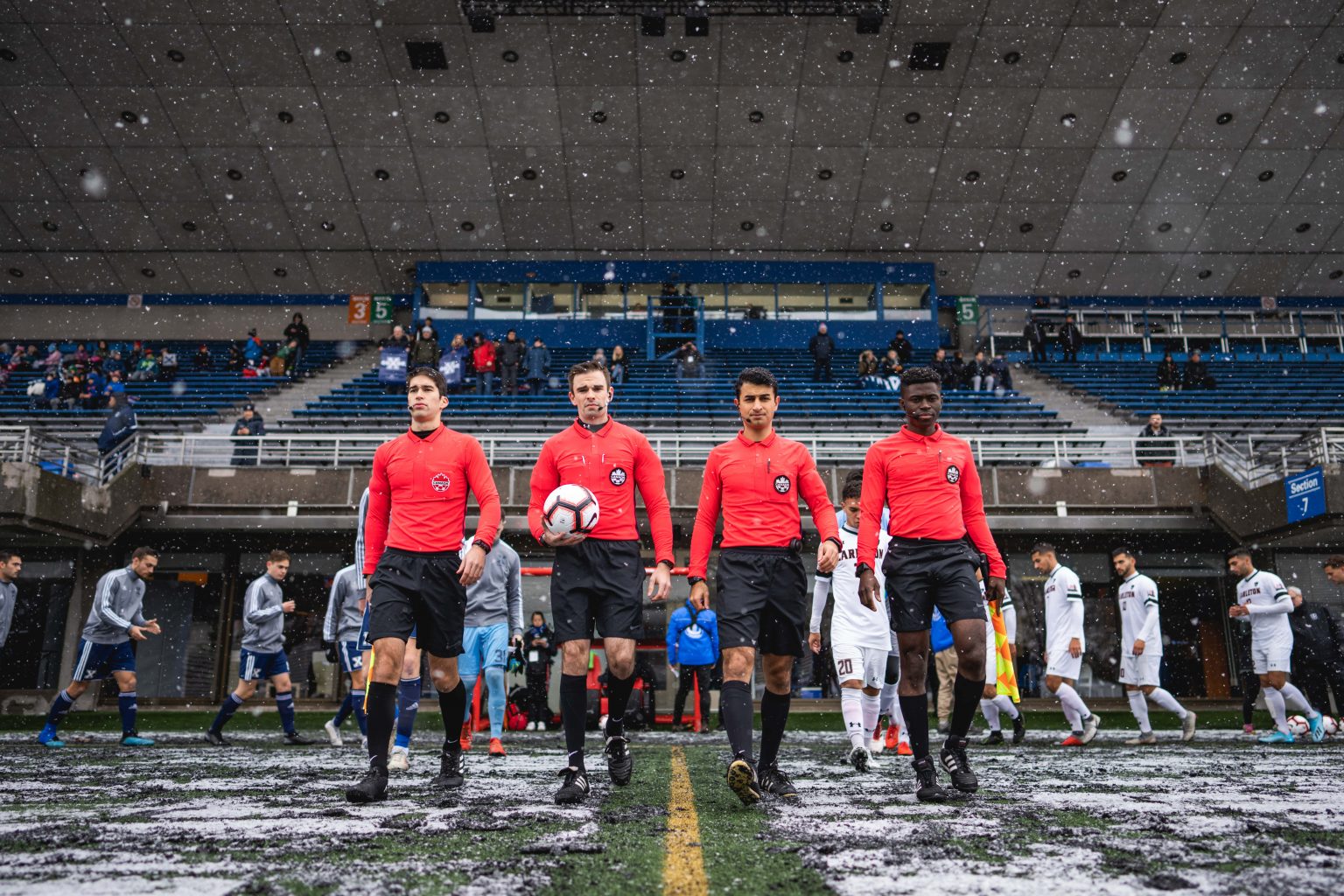Four soccer players enter a snowy stadium wearing red and black uniforms. 