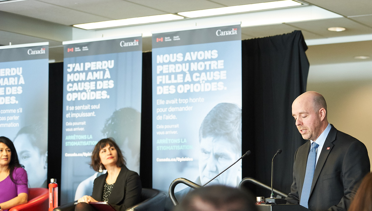 President Bacon speaks at a podium at an Opioid anti-stigma event. Prof. Sarah Everts and another panel guest listen on stage.