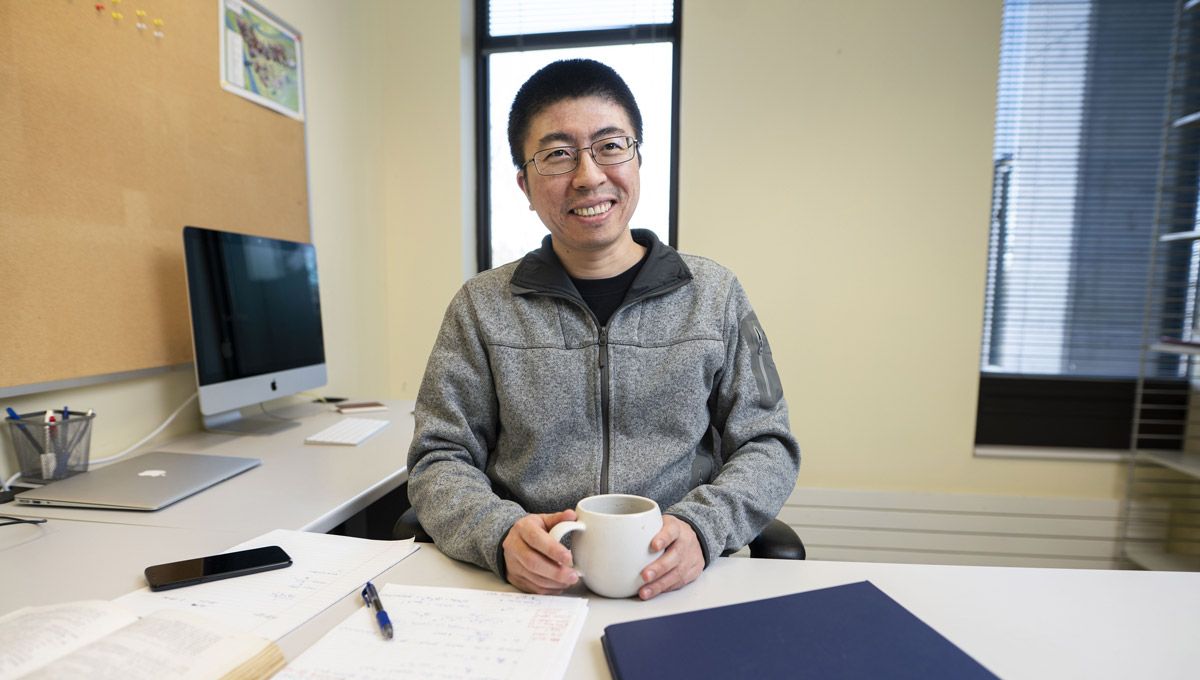 Yue Zhang sits at his desk with a Macbook, iMac, mobile phone in the background. He's holding a cup of coffee and smiling at the camera.