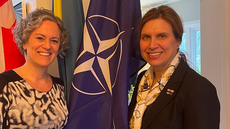 Two women - the ambassador of Canada to Austria, and the ambassador of Sweden to Austria - posing for a photo with various flags in between them.