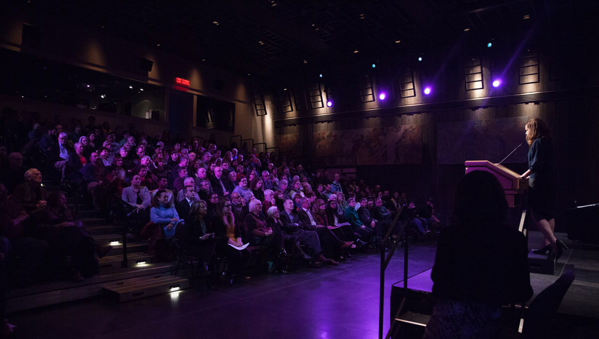 A crowd listens as Adrienne Arsenault is intruduces by a speaker at a podium during the Peter Stursberg Foreign Correspondents Lecture