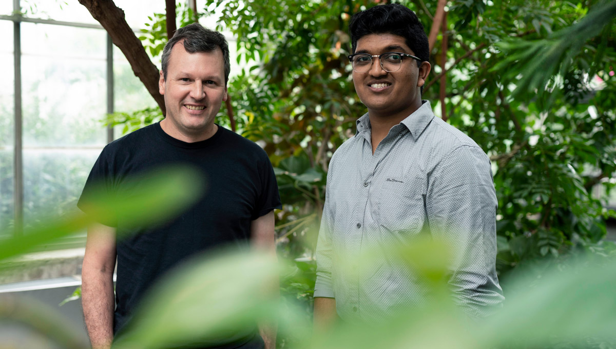 Prof. Bill Willmore and Bhavya Mohan pose in the greenhouse surrounded by plants.