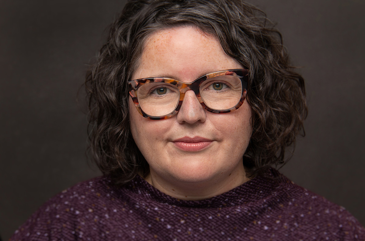 A professional headshot of woman wearing glasses and a purple sweater.