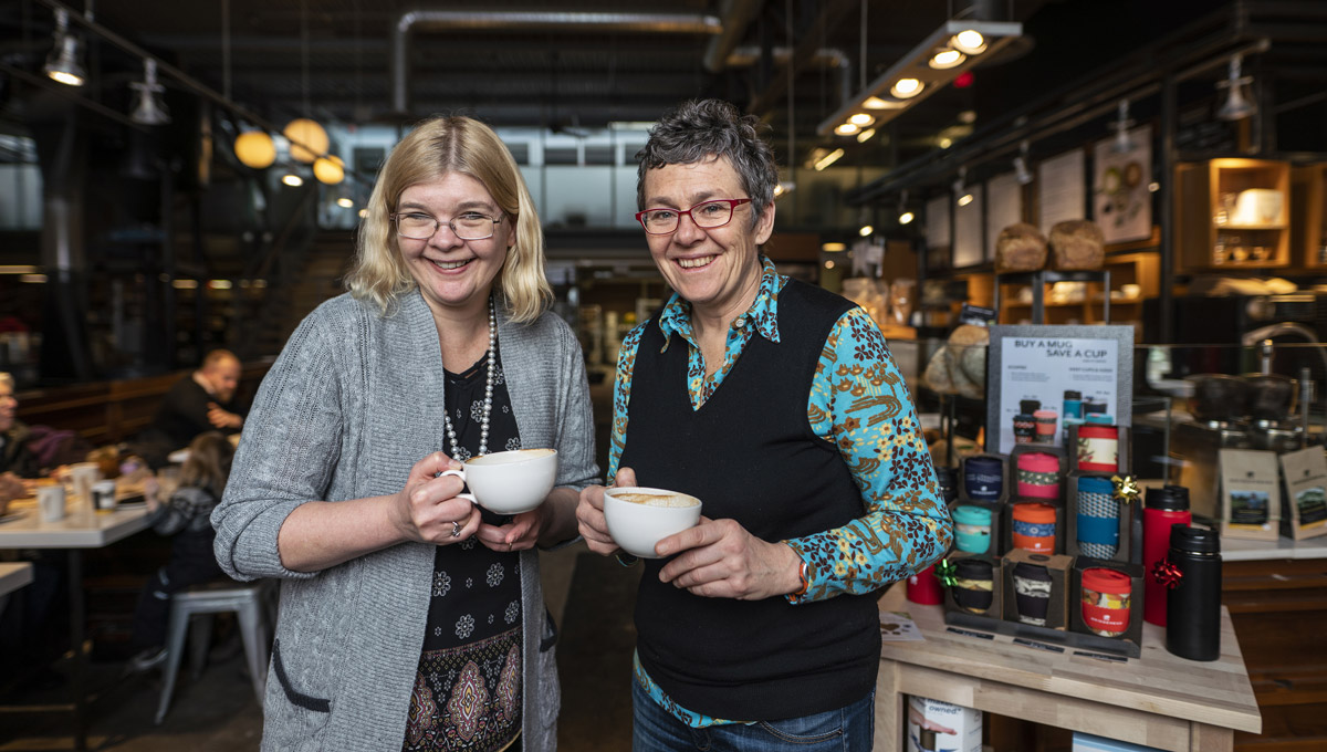 Tracey Clark and Dana Brown pose while holding cups of coffee at a Bridgehead.