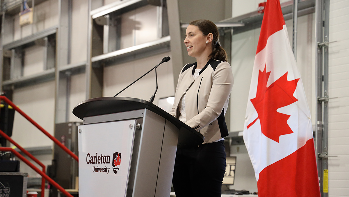 A woman in business suit speaks from a podium with a Canadian flag visible behind her.