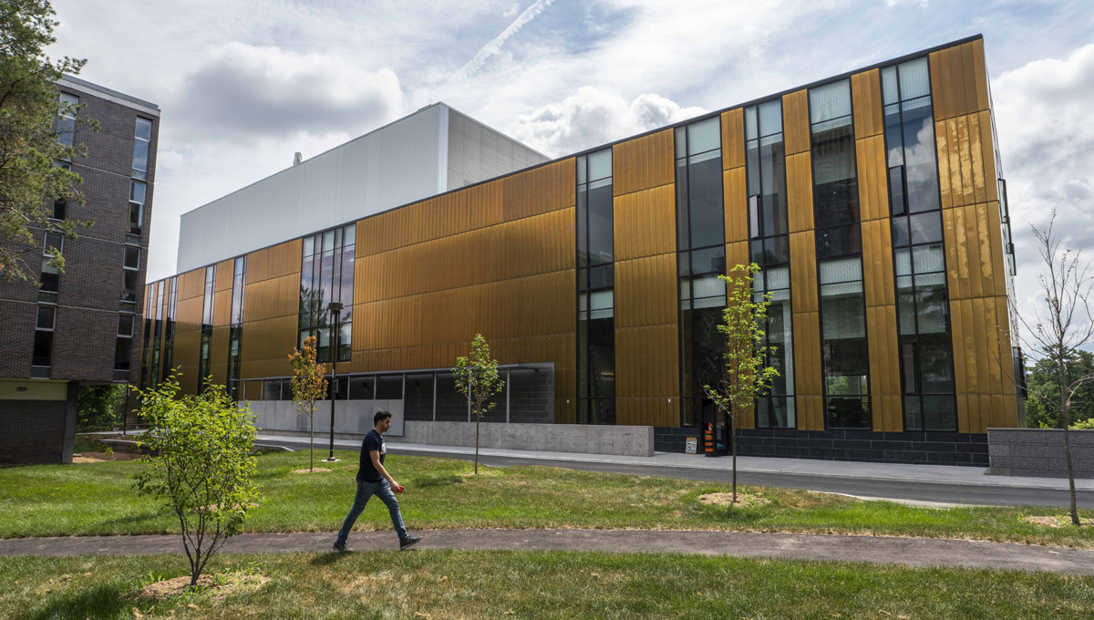 A student walks across the grass outside of the metal-plated ARISE Building on a sunny day.