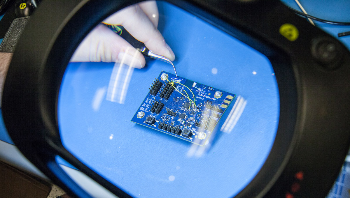 A closeup of a printed circuit board labeled with the Tweed branding as seen through a magnifying glass while a gloved hand solders it.