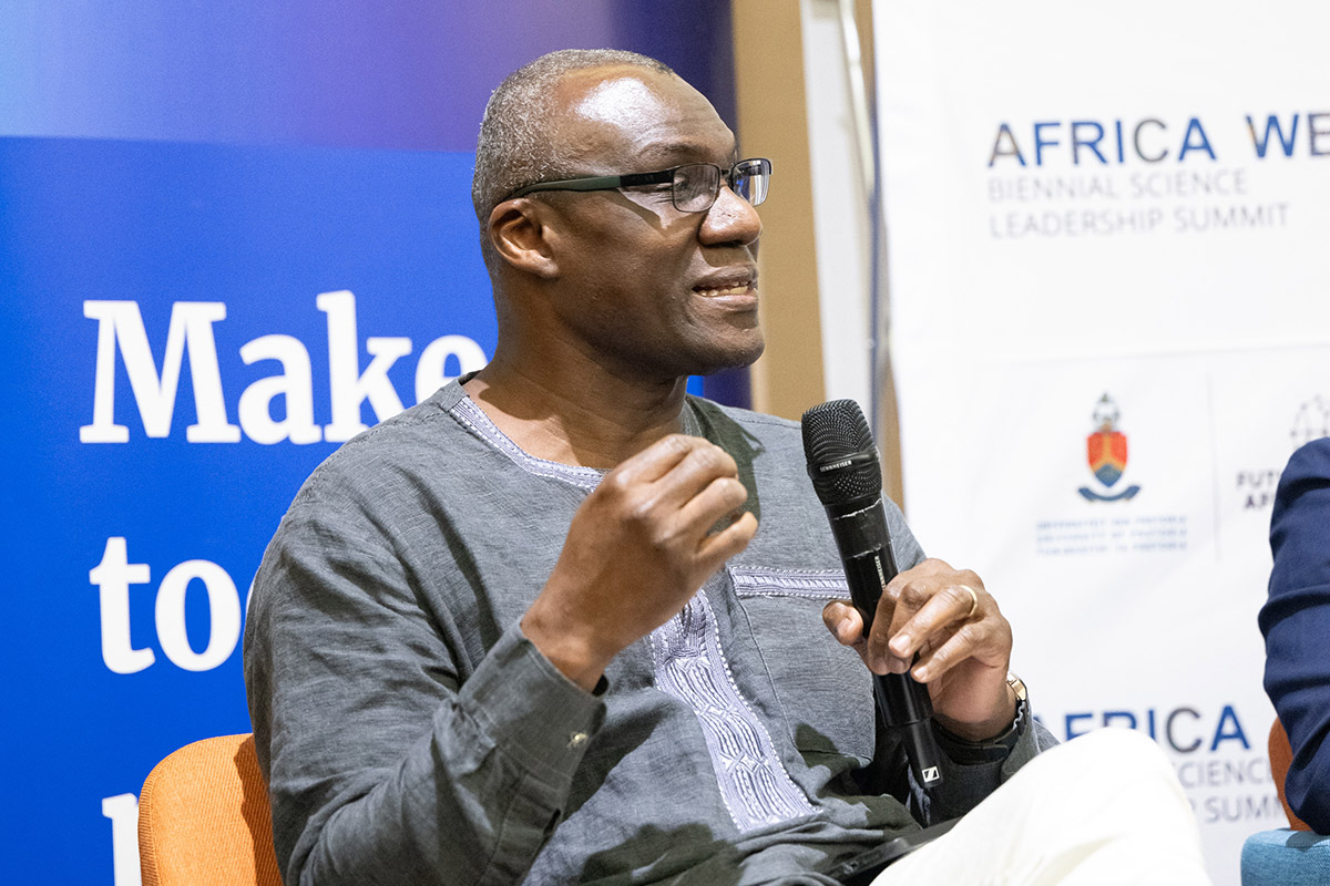 A seated man holding a microphone speaks to an audience.