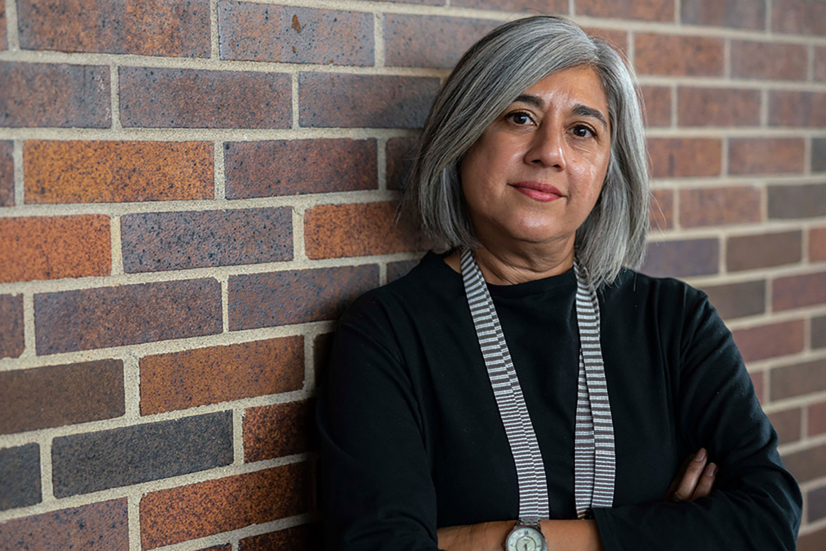 A woman with her arms crossed, leans against a brick wall to pose for a photo.