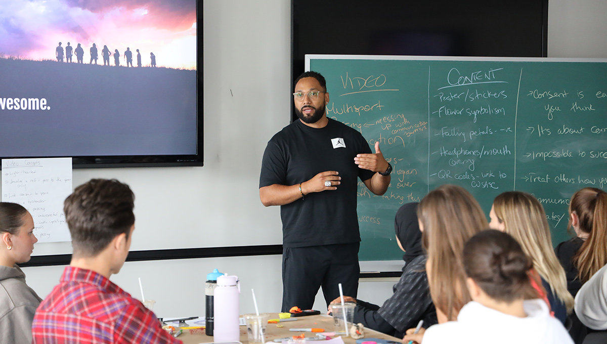 A man dressed in all black stands at the front of a classroom, addressing students during a Champions for Change session.